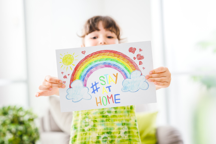 Child holding a drawing of a rainbow with the words 'stay at home' written in the middle