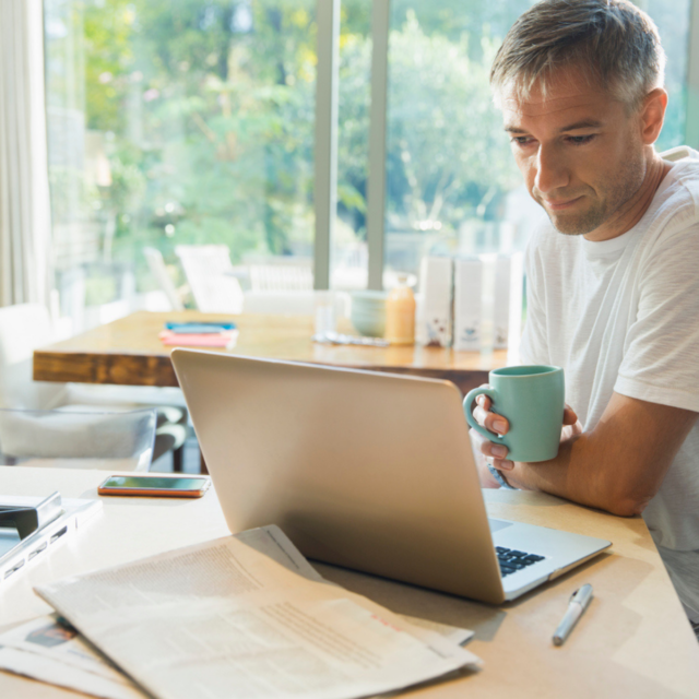 Man sat in kitchen, looking at laptop with bills around him - Household Spending Keeping Economy Afloat